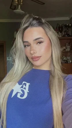 Long-haired blonde woman in a blue sweater takes a close-up selfie indoors.