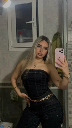 Blonde woman in a black strapless top and jeans taking a mirror selfie in a bathroom.