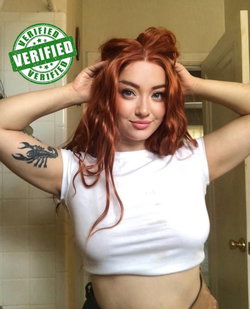 Redhead woman in white top with tattoo poses in bathroom.