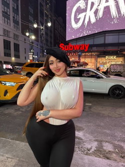 Curvy woman in a white sheer top and black leggings poses on a city street at night, wearing a black beret with tattoos