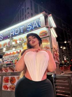 Curvy woman in a white corset top and black skirt poses confidently in front of a lit hot dog cart at night, wearing a