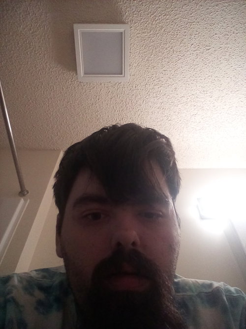 Bearded man with dark bangs takes bathroom selfie wearing tie-dye shirt.