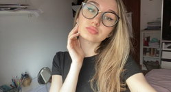 Blonde woman with glasses in a black t-shirt poses in her bedroom, hand near face, looking at the camera.