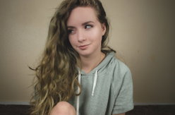 A young woman with long wavy blonde hair wears a gray hoodie while sitting indoors against a plain wall.