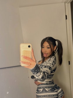 A woman with dark hair in a ponytail takes a mirror selfie, wearing a patterned long-sleeve top and skirt, sticking her