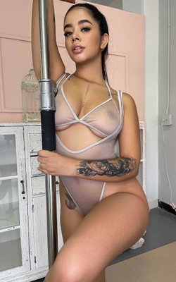 Curvy woman in sheer pink bodysuit with white straps poses by a pole, showcasing arm tattoos in a room with pink walls