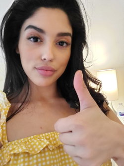 Woman with long dark hair in a yellow checkered top gives a thumbs-up in a softly lit room.