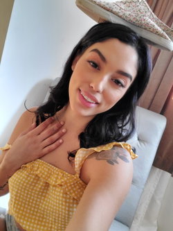A woman with dark hair and a nose ring wears a yellow checkered top, smiling while holding her chest; a floral sneaker