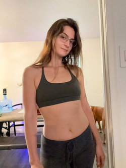A woman with long hair and glasses stands in a room wearing a black sports bra and leggings.