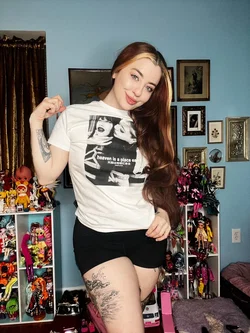 Smiling woman with long reddish hair and tattoos wears white graphic tee and black shorts in doll-filled room.