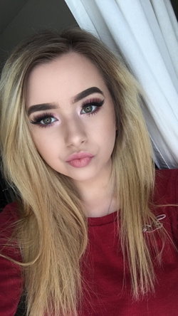 Blonde woman with long hair and makeup, wearing a red top, taking a close-up selfie indoors near a white curtain.