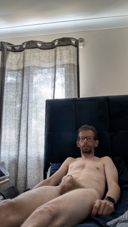 A shirtless man with glasses and a beard lies on a bed with a dark headboard, partially covered by sheets, in a room