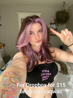 A young woman with purple-tipped hair and colorful arm tattoos takes a selfie in a bedroom, wearing a black crop top.