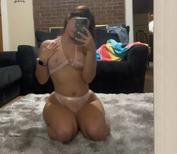 Woman in pink lace lingerie kneeling on a fluffy rug, taking a mirror selfie in a living room with a couch and brick