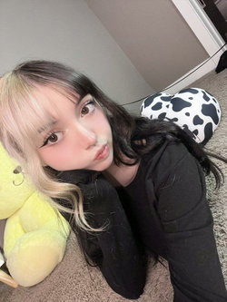 Woman with black-and-blonde split hair, lip piercing, pouting in black hoodie on floor with stuffed animals.