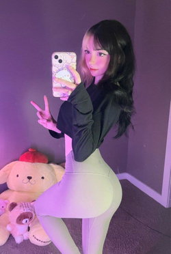 Woman with pink-and-black split hair poses peace sign selfie in black crop top and tight gray leggings in bedroom with