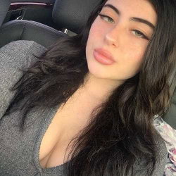 Woman with long dark hair in a car, wearing a gray top.