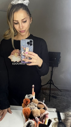 Blonde woman in black sweatshirt takes mirror selfie, holding phone with decorated case, makeup brushes in foreground.