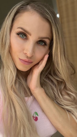 Blonde woman with long wavy hair and green eyes, wearing a pink cherry-embroidered top, posing indoors with hand on