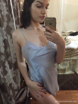 Brunette woman in a light blue lace-trimmed camisole takes a mirror selfie in a bedroom, hand resting on her hip.