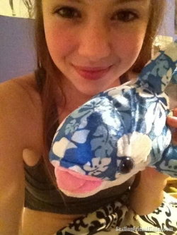 A smiling woman with long brown hair holds a blue floral plush dolphin, wearing a dark crop top and patterned bottoms