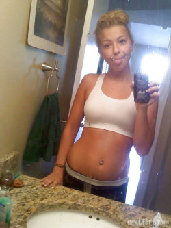 Blonde woman in white sports bra and jeans takes mirror selfie in bathroom, holding a drink, with visible belly button