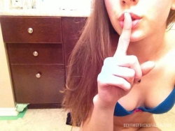 Brunette woman in blue bra holding finger to lips, selfie-style shot in bathroom setting.
