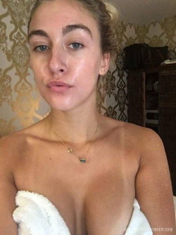 Blonde woman with freckles wrapped in a white towel, bare shoulders, gold necklace, patterned wallpaper background.