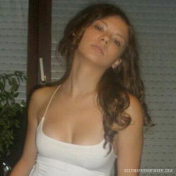 Brunette woman with wavy hair wearing a white tank top, standing indoors near a window with blinds and a plant.