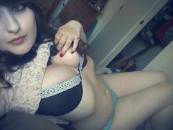 A woman with dark hair in a lace top and black bra, lying down in a bedroom setting with visible lingerie.