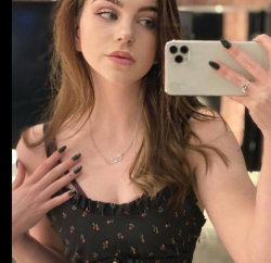 A woman with long brown hair takes a mirror selfie, wearing a black floral top and dark nail polish, hand resting on