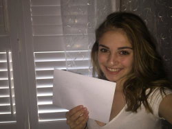A smiling woman with shoulder-length brown hair wears a white top and holds a piece of paper in front of a window with