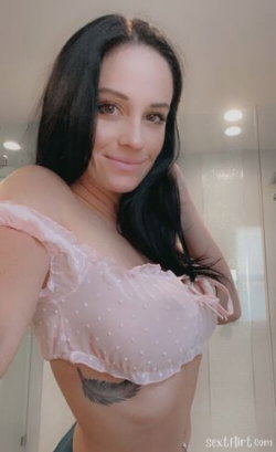 Brunette woman with long dark hair in a sheer pink top, smiling at the camera in a bathroom setting.