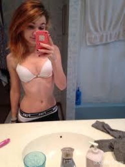 A woman with shoulder-length reddish hair takes a mirror selfie in a bathroom, wearing a white bra and black underwear