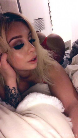 Blonde woman with tattoos resting on a bed, partially covered by a white fur blanket, in a dimly lit bedroom setting.