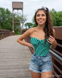 A smiling woman with long dark hair stands on a wooden bridge, wearing a green striped strapless top and denim shorts.