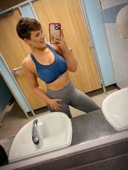 A woman with short hair in a blue sports bra and gray leggings takes a mirror selfie in a public bathroom.