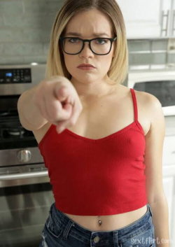 A blonde woman in a red crop top and jeans points directly at the camera in a modern kitchen.