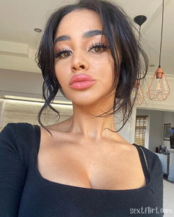 A woman with dark hair and makeup wears a black top in a modern indoor setting, taking a close-up selfie.