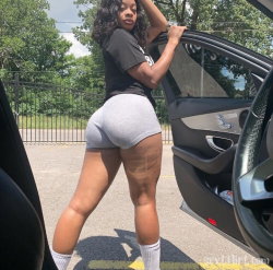 Curvy woman in black t-shirt and gray shorts leaning against open car door outdoors.