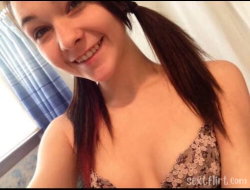 A smiling woman with dark hair in a ponytail wearing a floral-patterned bra, taking a close-up selfie indoors near a