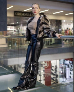A woman with blonde hair and red lipstick wears a beige top, black leather pants, and a fur-trimmed jacket while