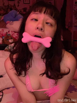 A young woman with dark hair and a nose ring sticks out her tongue while holding a pink bone-shaped toy in her mouth,