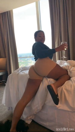 Curvy woman in a dark top and beige shorts poses on a bed holding a wine glass, with city view through large window