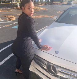 A curvy woman in a black textured bodysuit and heels poses by a white Mercedes in a parking lot, looking over her