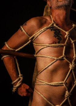 A muscular man with visible tattoos is bound with rope in a studio setting, shirtless and posing against a dark