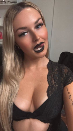 Blonde woman with dark lipstick and a nose ring, wearing a black lace top, posing indoors in a selfie-style shot.