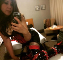 Brunette woman in black and red lingerie with fishnet stockings and high-heeled boots takes a mirror selfie in a hotel