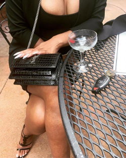 Curvy woman in black top and blazer sits at outdoor table, holding a textured clutch, with a cocktail glass and car