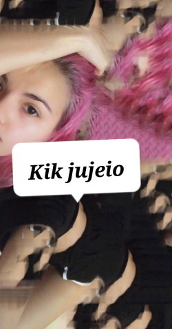 A woman with pink hair lies on a fuzzy pink surface, wearing black clothing, in a close-up selfie-style shot.